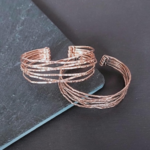 Peut inclure: Deux bracelets en m&eacute;tal couleur cuivre. L'un est de style manchette, l'autre est un jonc. Les deux ont un aspect textur&eacute; et martel&eacute;. Le bracelet manchette est compos&eacute; de plusieurs brins de m&eacute;tal. Le fond est une surface gris fonc&eacute; et noire.