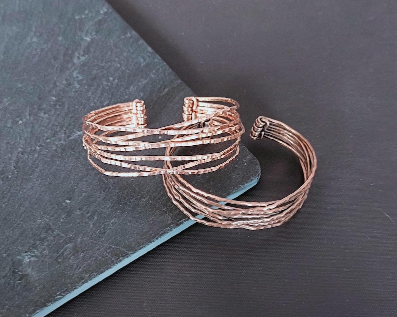 Peut inclure: Deux bracelets en m&eacute;tal couleur cuivre. L'un est de style manchette, l'autre est un jonc. Les deux ont un aspect textur&eacute; et martel&eacute;. Le bracelet manchette est compos&eacute; de plusieurs brins de m&eacute;tal. Le fond est une surface gris fonc&eacute; et noire.