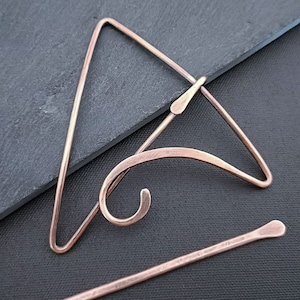 May include: A copper hair pin with a triangular shape and a curved design. The pin is on a dark grey surface.