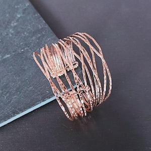 Peut inclure: Un bracelet manchette de couleur cuivr&eacute;e avec une finition martel&eacute;e textur&eacute;e. Le bracelet est compos&eacute; de plusieurs brins de m&eacute;tal fins et incurv&eacute;s, cr&eacute;ant un design ouvert et superpos&eacute;. Le bijou est pr&eacute;sent&eacute; sur une surface gris fonc&eacute;.