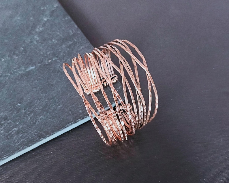 Peut inclure: Un bracelet manchette de couleur cuivr&eacute;e avec une finition martel&eacute;e textur&eacute;e. Le bracelet est compos&eacute; de plusieurs brins de m&eacute;tal fins et incurv&eacute;s, cr&eacute;ant un design ouvert et superpos&eacute;. Le bijou est pr&eacute;sent&eacute; sur une surface gris fonc&eacute;.