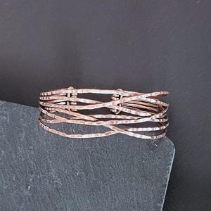 Peut inclure: Une bague de couleur cuivr&eacute;e avec un design textur&eacute; et martel&eacute;. La bague est compos&eacute;e de plusieurs brins de fil, tiss&eacute;s ensemble pour cr&eacute;er un effet superpos&eacute; unique. La bague est pr&eacute;sent&eacute;e sur une surface gris fonc&eacute;.