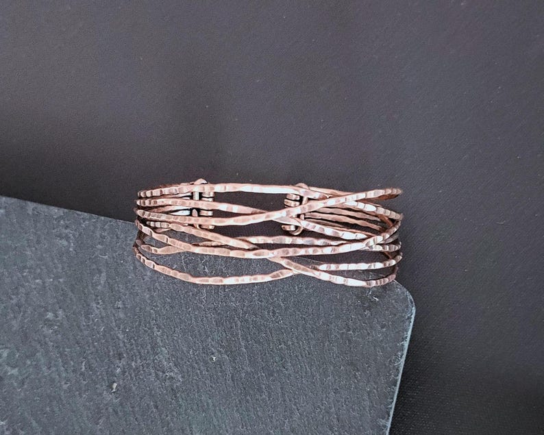 Peut inclure: Une bague de couleur cuivr&eacute;e avec un design textur&eacute; et martel&eacute;. La bague est compos&eacute;e de plusieurs brins de fil, tiss&eacute;s ensemble pour cr&eacute;er un effet superpos&eacute; unique. La bague est pr&eacute;sent&eacute;e sur une surface gris fonc&eacute;.