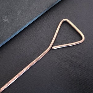 May include: A long, thin, copper-coloured metal rod with a triangular loop at one end. The rod has a hammered texture.