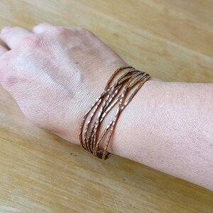 Peut inclure: Un ensemble de bracelets de couleur cuivre avec une texture martel&eacute;e. Les bracelets sont port&eacute;s sur un poignet, et le fond est une surface en bois de couleur claire.