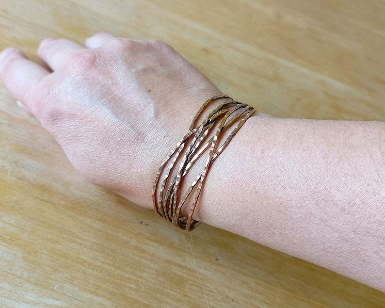 Peut inclure: Un ensemble de bracelets de couleur cuivre avec une texture martel&eacute;e. Les bracelets sont port&eacute;s sur un poignet, et le fond est une surface en bois de couleur claire.