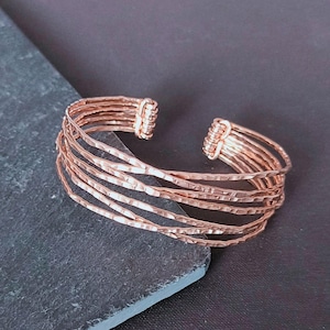 Peut inclure: Bracelet manchette de couleur cuivre avec une texture martel&eacute;e. Le design ouvert pr&eacute;sente plusieurs bandes parall&egrave;les, cr&eacute;ant un effet superpos&eacute;. Les extr&eacute;mit&eacute;s sont envelopp&eacute;es de fil. Un accessoire &eacute;l&eacute;gant.