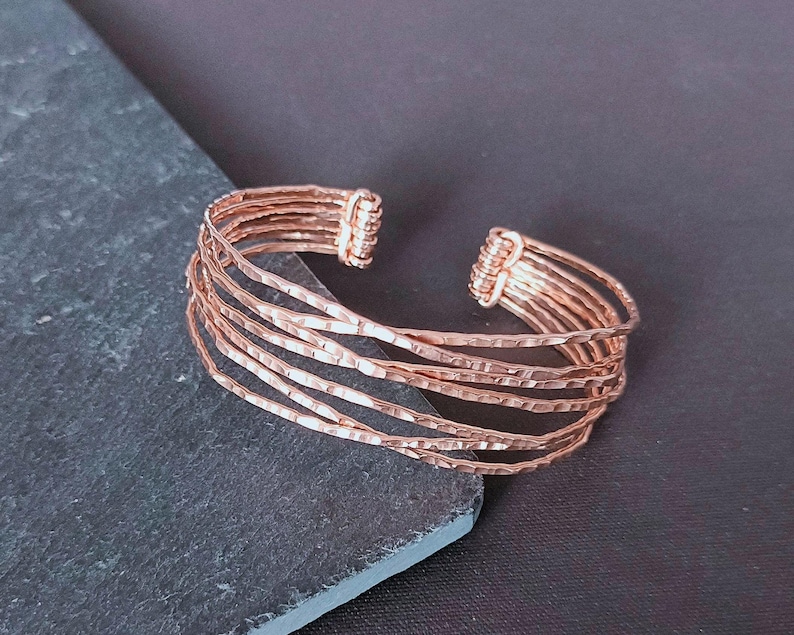Peut inclure: Bracelet manchette de couleur cuivre avec une texture martel&eacute;e. Le design ouvert pr&eacute;sente plusieurs bandes parall&egrave;les, cr&eacute;ant un effet superpos&eacute;. Les extr&eacute;mit&eacute;s sont envelopp&eacute;es de fil. Un accessoire &eacute;l&eacute;gant.