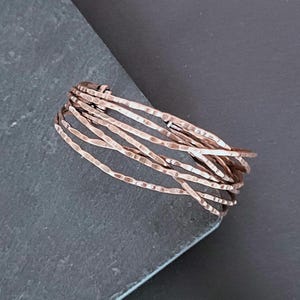 May include: A collection of hammered copper rings. The rings have a textured, uneven surface and are grouped together. The copper has a warm, reddish-brown colour, and the rings are shown on a dark grey surface.