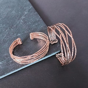 Peut inclure: Deux bracelets de manchette en m&eacute;tal couleur cuivre. L'un des bracelets a plusieurs bandes parall&egrave;les, tandis que l'autre a un design plus ouvert avec des barres verticales. Les bracelets sont pr&eacute;sent&eacute;s sur une surface gris fonc&eacute; sur fond noir.