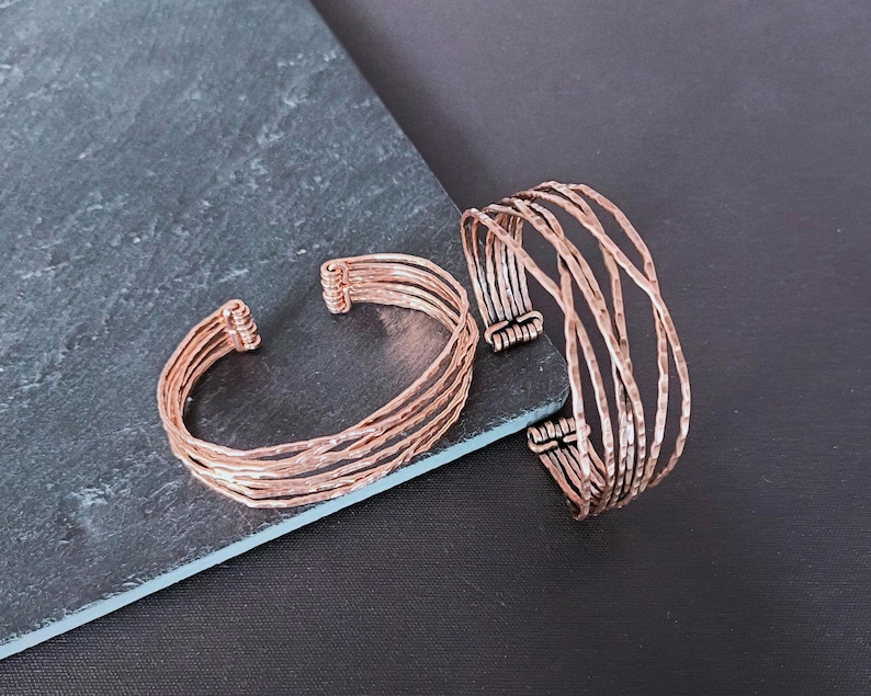 Peut inclure: Deux bracelets de manchette en m&eacute;tal couleur cuivre. L'un des bracelets a plusieurs bandes parall&egrave;les, tandis que l'autre a un design plus ouvert avec des barres verticales. Les bracelets sont pr&eacute;sent&eacute;s sur une surface gris fonc&eacute; sur fond noir.