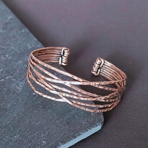 Peut inclure: Bracelet manchette en cuivre &agrave; texture martel&eacute;e. Le bracelet ouvert pr&eacute;sente plusieurs bandes parall&egrave;les qui se croisent. Le m&eacute;tal a une teinte brun-rouge&acirc;tre chaude, et le design est simple mais saisissant.
