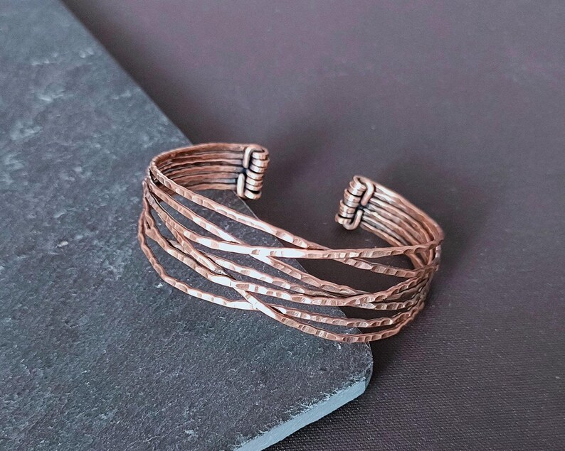 Peut inclure: Bracelet manchette en cuivre &agrave; texture martel&eacute;e. Le bracelet ouvert pr&eacute;sente plusieurs bandes parall&egrave;les qui se croisent. Le m&eacute;tal a une teinte brun-rouge&acirc;tre chaude, et le design est simple mais saisissant.