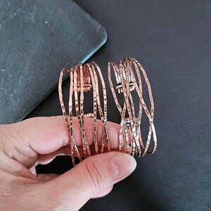 Peut inclure: Deux bracelets de couleur cuivre avec une texture martel&eacute;e. Les bracelets sont compos&eacute;s de plusieurs brins fins et verticaux, cr&eacute;ant un design ouvert et a&eacute;r&eacute;. Les bracelets sont pr&eacute;sent&eacute;s sur un fond sombre.