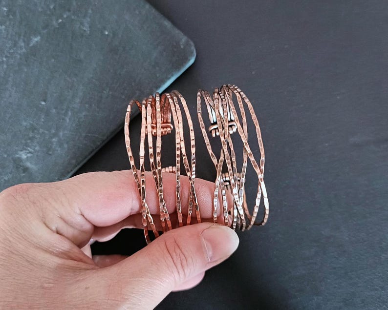 Peut inclure: Deux bracelets de couleur cuivre avec une texture martel&eacute;e. Les bracelets sont compos&eacute;s de plusieurs brins fins et verticaux, cr&eacute;ant un design ouvert et a&eacute;r&eacute;. Les bracelets sont pr&eacute;sent&eacute;s sur un fond sombre.