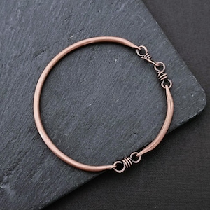 Può includere: Un braccialetto a cerchio color rame dal design semplice e minimalista. Il braccialetto è rotondo con una chiusura a fermaglio. Il metallo ha una calda tonalità marrone-rossastra. Il braccialetto è mostrato su una superficie grigio scuro.