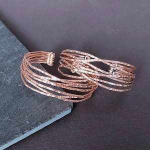 Peut inclure: Deux bracelets manchettes ouverts de couleur cuivr&eacute;e. Chaque bracelet est compos&eacute; de plusieurs fines brins de m&eacute;tal martel&eacute;. Les bracelets sont pr&eacute;sent&eacute;s sur une surface grise fonc&eacute;e et noire, cr&eacute;ant un contraste qui met en valeur la texture et le design des bijoux.