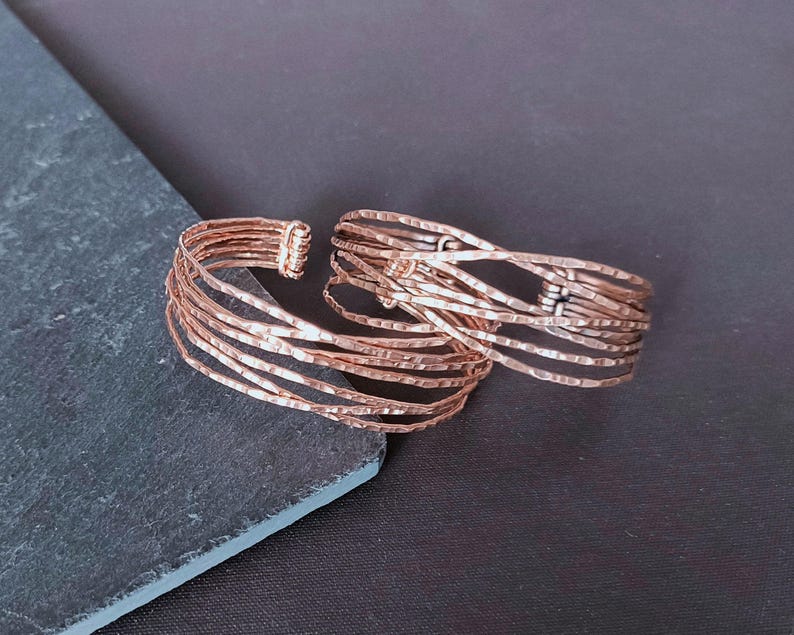 Peut inclure: Deux bracelets manchettes ouverts de couleur cuivr&eacute;e. Chaque bracelet est compos&eacute; de plusieurs fines brins de m&eacute;tal martel&eacute;. Les bracelets sont pr&eacute;sent&eacute;s sur une surface grise fonc&eacute;e et noire, cr&eacute;ant un contraste qui met en valeur la texture et le design des bijoux.