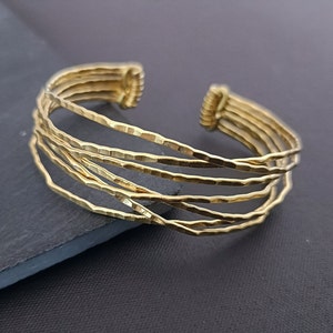 Może przedstawiać: Złota bransoletka typu bangle wykonana z wielu cienkich, młotkowanych pasków metalu. Bransoletka jest otwarta i regulowana.