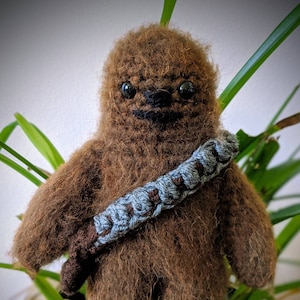 Puede incluir: Un juguete de peluche Chewbacca de crochet marrón con un bandolera gris y marrón. El juguete está sentado en una planta verde.