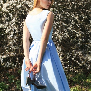 May include: A woman in a light blue dress stands in front of a white flowering bush. She is holding a black shoe in her hand.
