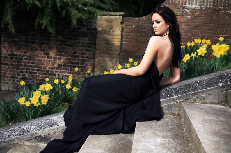 May include: A woman in a black dress poses on a set of stone steps with a background of yellow flowers and a brick wall.