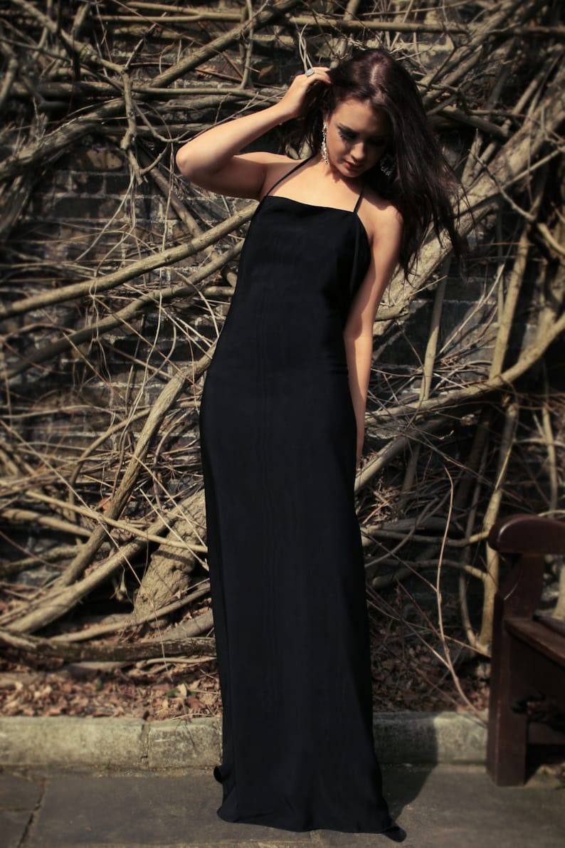 May include: A woman wearing a long black dress stands in front of a wall of branches. She is looking down and has her hand on her ear.