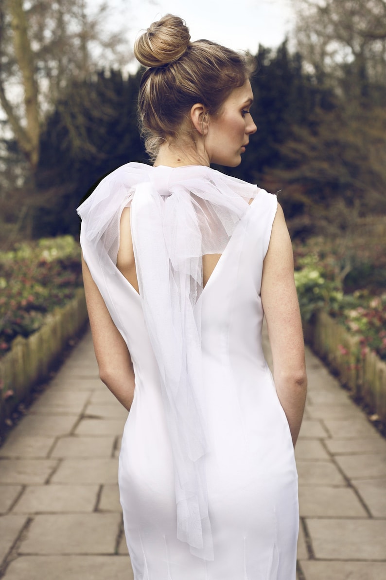 May include: A white sleeveless dress with a large white bow at the back of the neck. The dress is fitted and has a simple design.