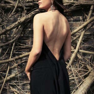 May include: A woman wearing a black backless dress with a low neckline. She is standing in front of a background of brown branches.