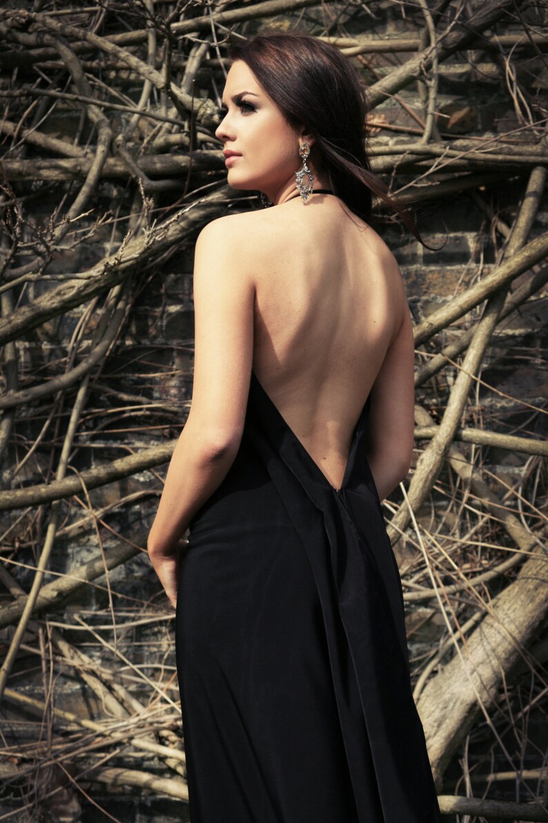 May include: A woman wearing a black backless dress with a low neckline. She is standing in front of a background of brown branches.