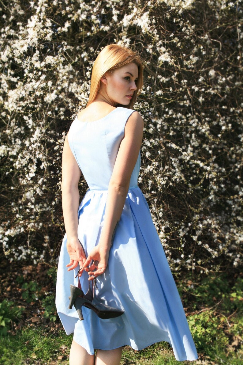 May include: A woman in a light blue dress stands in front of a white flowering bush. She is holding a black shoe in her hand.
