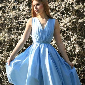 May include: A woman wearing a light blue sleeveless dress with a V-neckline and a gathered skirt. She is standing in front of a white flowering bush.