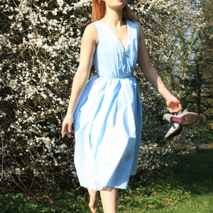 May include: A woman in a light blue dress walks barefoot through a field of white flowers. She is smiling and holding a pair of shoes in her hand.