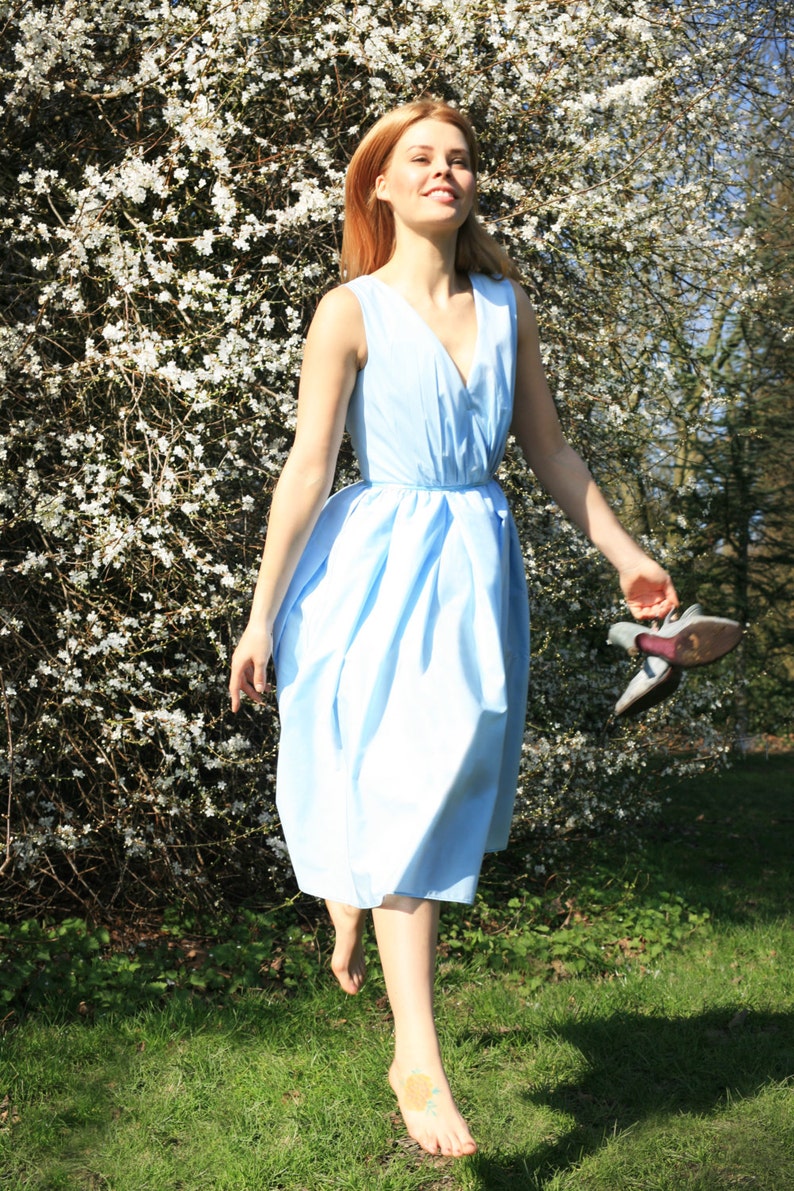 May include: A woman in a light blue dress walks barefoot through a field of white flowers. She is smiling and holding a pair of shoes in her hand.