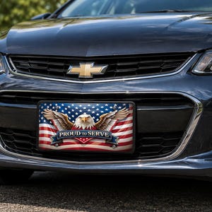 May include: A rectangular license plate frame featuring a graphic of a bald eagle with wings spread wide against a backdrop of the American flag. A blue banner across the bottom displays the white text PROUD TO SERVE in a bold, capitalized font.