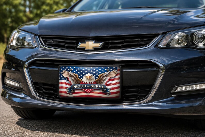 May include: A rectangular license plate frame featuring a graphic of a bald eagle with wings spread wide against a backdrop of the American flag. A blue banner across the bottom displays the white text PROUD TO SERVE in a bold, capitalized font.