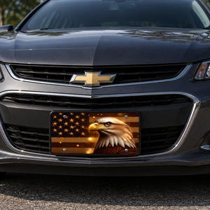 May include: A dark gray Chevrolet sedan featuring a front license plate with a graphic of a bald eagle head set against a stylized American flag background with golden stars and horizontal stripes, mounted on the front bumper grille.