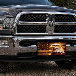 May include: A dark gray pickup truck with a chrome grille and bumper featuring a rectangular license plate. The plate displays a graphic of a bald eagle head set against a stylized American flag with warm golden and brown tones.