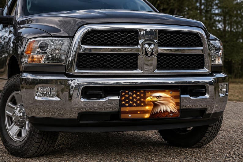 May include: A dark gray pickup truck with a chrome grille and bumper featuring a rectangular license plate. The plate displays a graphic of a bald eagle head set against a stylized American flag with warm golden and brown tones.