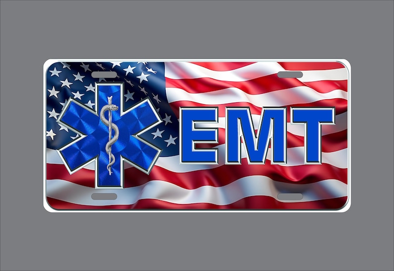 May include: A rectangular license plate featuring a waving American flag background. On the left is a blue Star of Life symbol with a silver serpent and staff. To the right, the letters EMT are printed in bold blue with a silver outline.