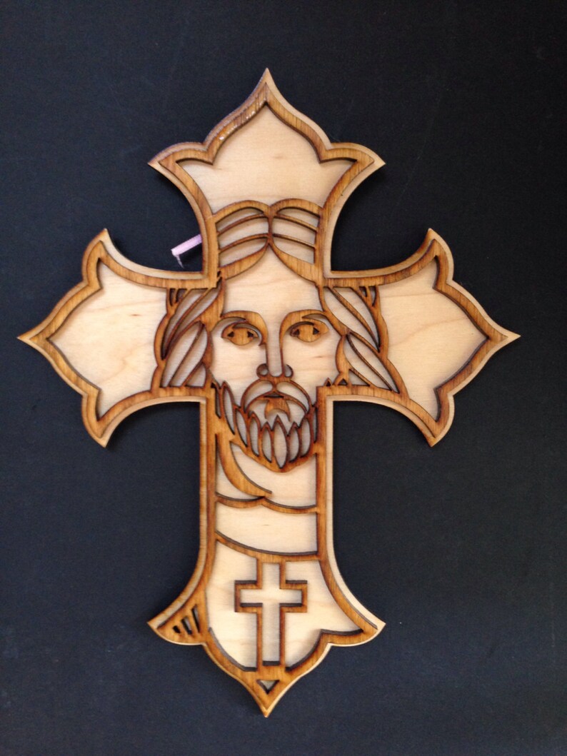 May include: A wooden cross with a detailed carving of Jesus' face in the center. The cross is made of light wood and has a brown stain.