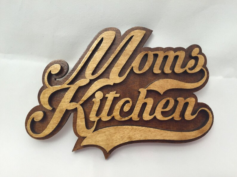 May include: A wooden sign with the words "Mom's Kitchen" in a decorative script font. The sign is stained in a dark brown color with a lighter brown wood grain visible.