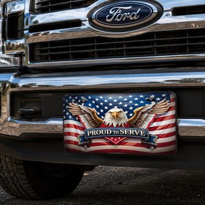 May include: A rectangular license plate mounted on the front bumper of a Ford truck. The plate features a graphic of the American flag, a bald eagle with wings spread, and a blue banner with the white text PROUD TO SERVE centered on the front.