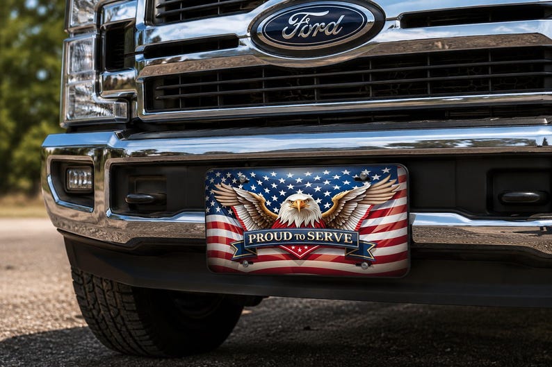 May include: A rectangular license plate mounted on the front bumper of a Ford truck. The plate features a graphic of the American flag, a bald eagle with wings spread, and a blue banner with the white text PROUD TO SERVE centered on the front.
