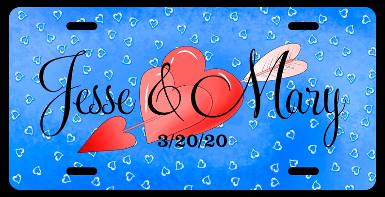 May include: A blue license plate with a red heart pierced by an arrow with white feathers. The text "Jesse & Mary 3/20/20" is written in black cursive script on the plate. The background is covered in small white hearts.