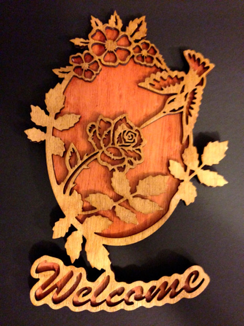 May include: Wooden welcome sign with a hummingbird and roses carved into the design. The sign is painted in shades of brown and orange.