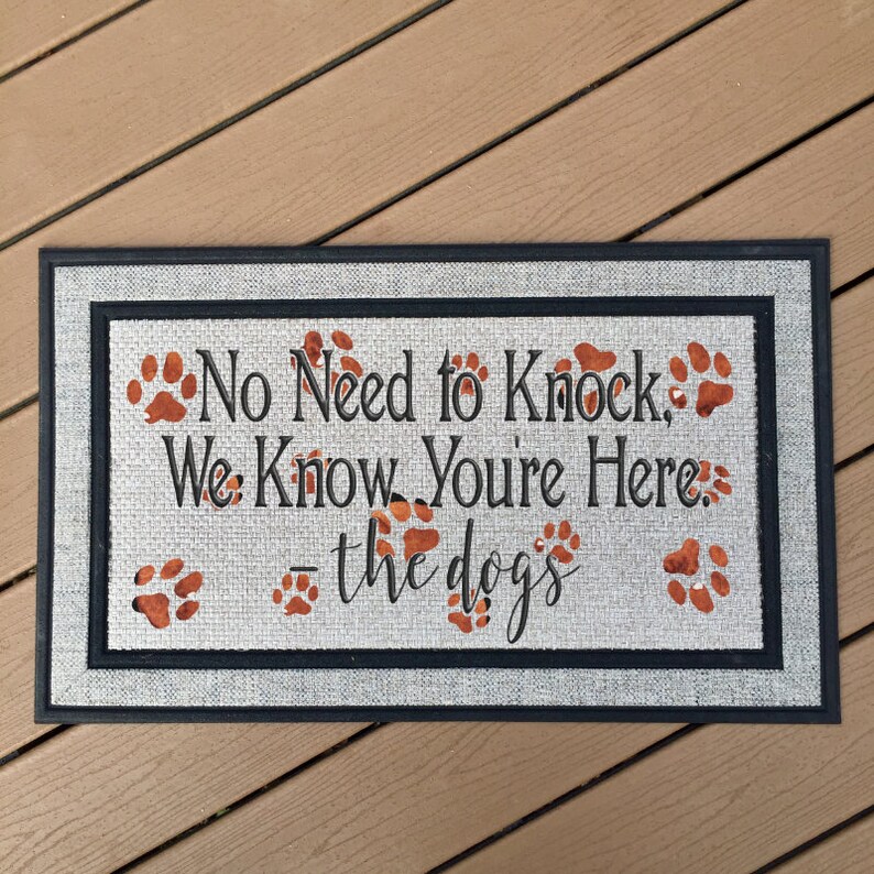 May include: A gray doormat with black trim and the text "No Need to Knock, We Know You're Here. - the dogs" in black. The doormat has several brown paw prints.