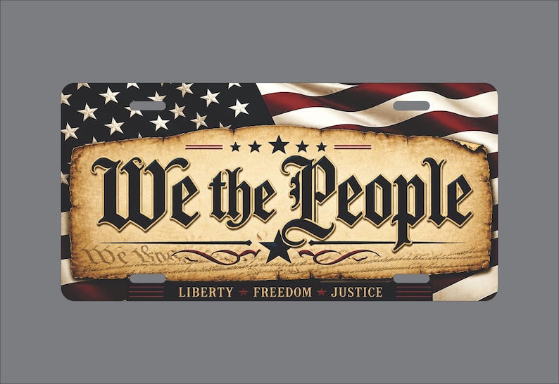 May include: A rectangular license plate featuring a waving American flag background. The center displays a parchment-style graphic with the black text We the People, a central star, and the words Liberty, Freedom, and Justice at the bottom.