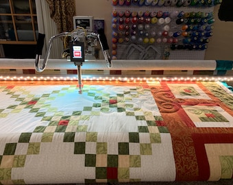 May include: A longarm quilting machine with a white quilt on the bed. The quilt has a patchwork design with green, red, and white squares. The machine is equipped with a light bar and a needle that is moving across the quilt.