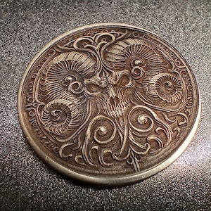 Puede incluir: Una moneda de plata con un diseño intrincado que presenta una calavera con cuernos de carnero y patrones en espiral.